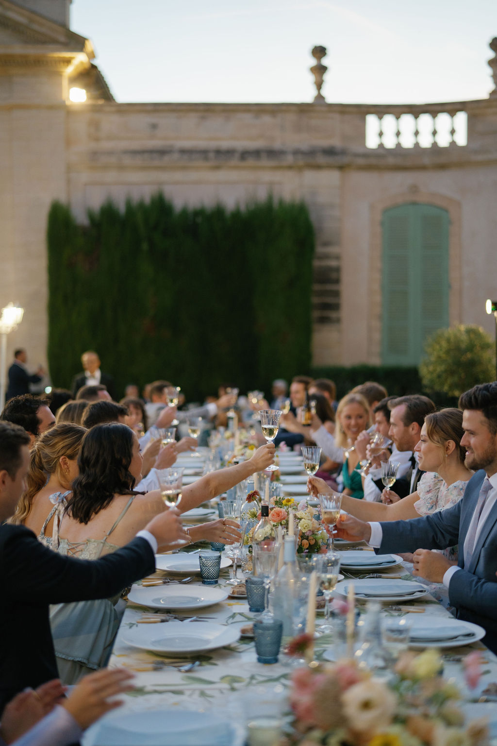 Guests gathered for an outdoor dinner at a Provence wedding, raising glasses at a long table with floral details and candlelight, planned by a Provence wedding planner.