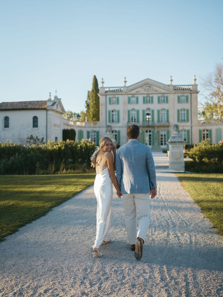 Couple walking along a gravel path toward a French chateau in Provence, part of an elegant destination wedding experience planned by a wedding planner France.