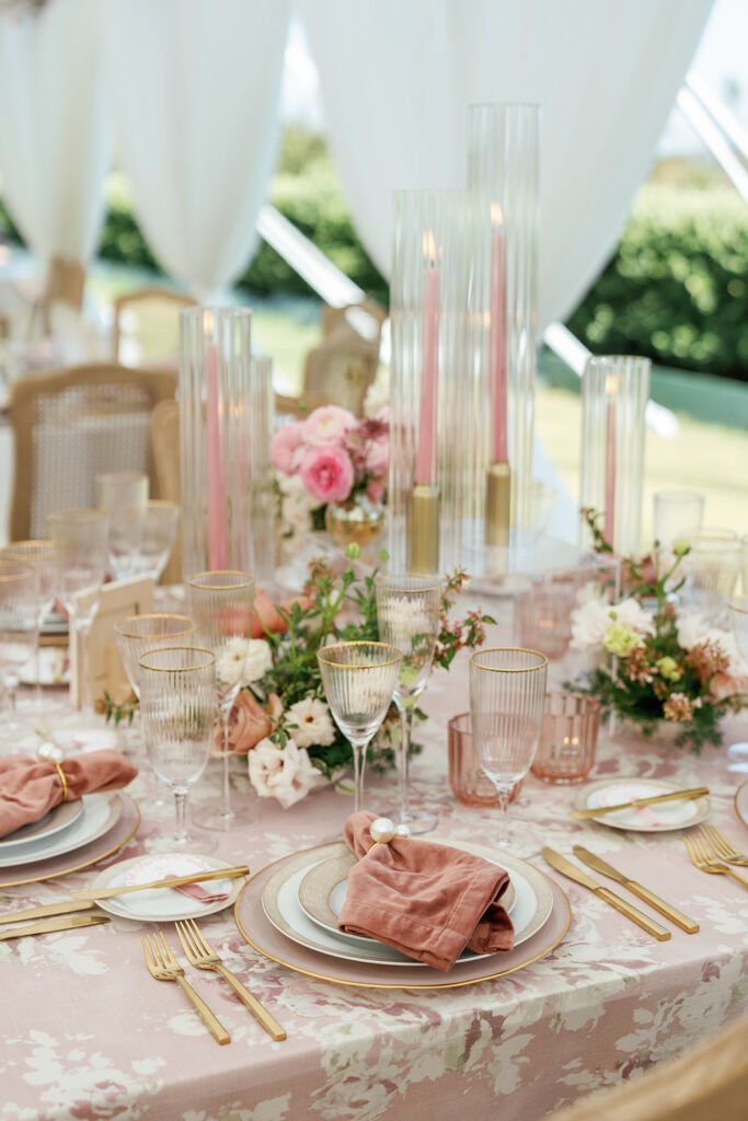 Elegant reception table with blush patterned linens, gold flatware, layered place settings, and soft pink floral arrangements.