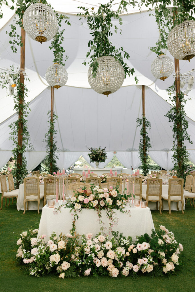 Sweetheart reception tables under a sailcloth tent with chandeliers, cascading greenery, and abundant floral centerpieces.