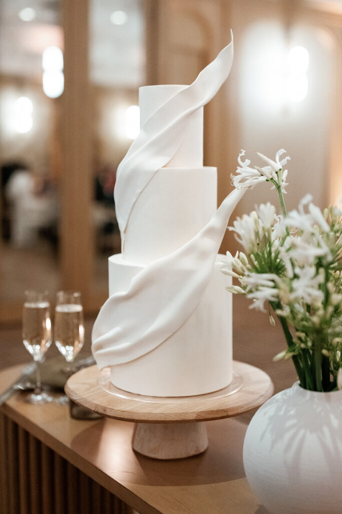 Modern sculptural wedding cake displayed on a custom stand with floral accents, representing design coordination included in full service wedding planning.