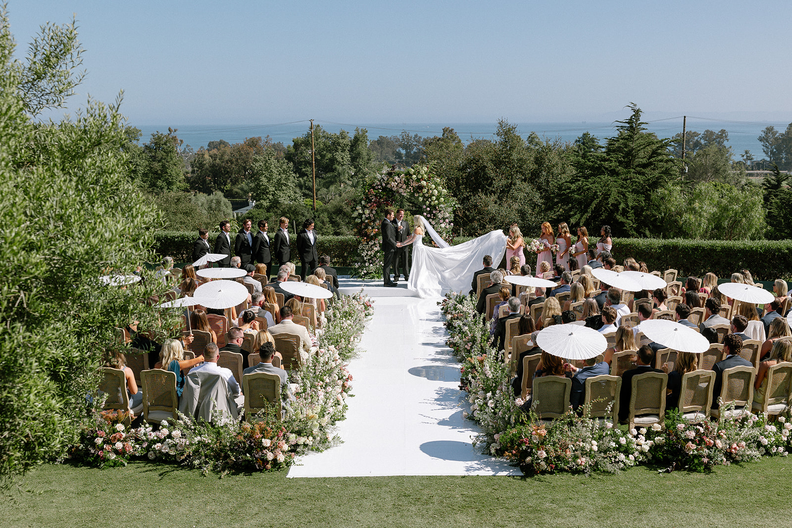 Outdoor California destination wedding ceremony with guests seated along a floral aisle and coastal views beyond.