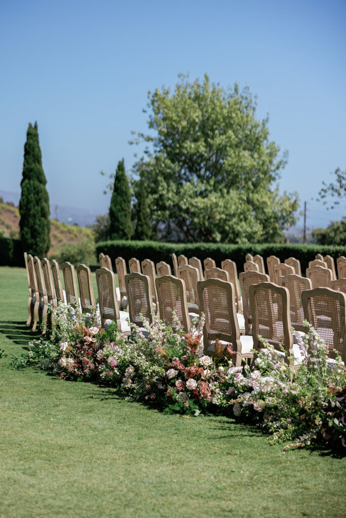 Outdoor ceremony seating with cane-back chairs arranged in rows, grounded by lush floral arrangements along the aisle edge.