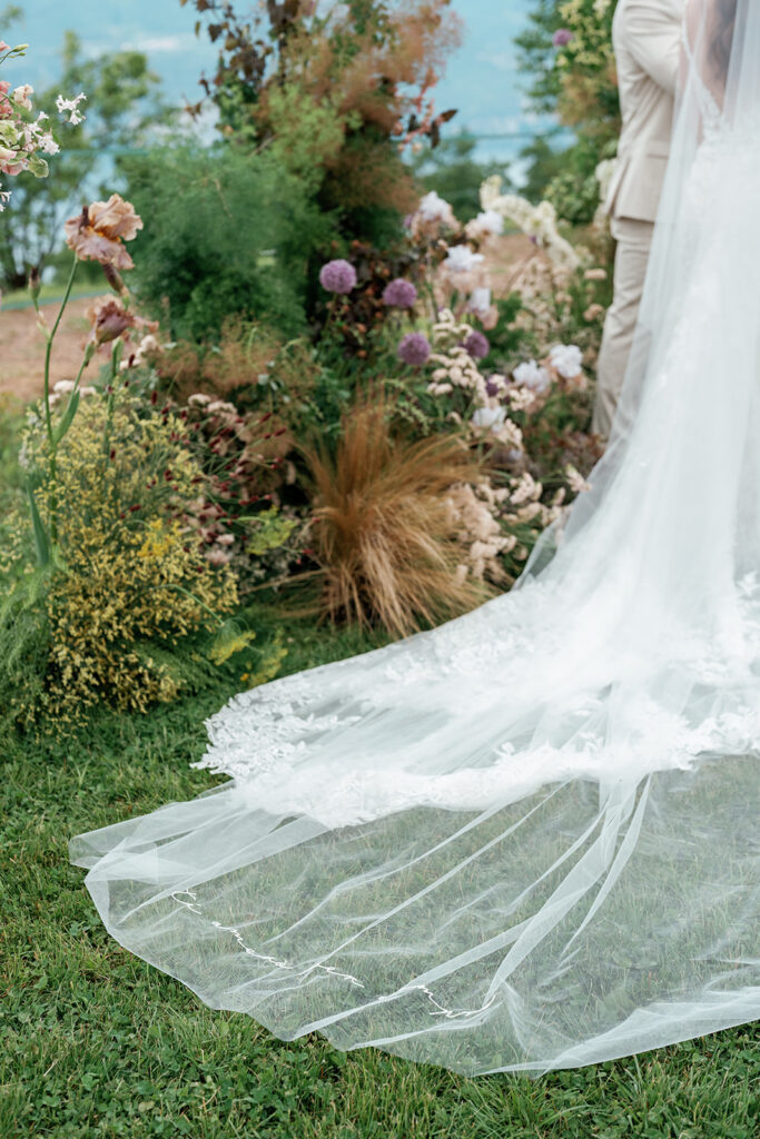 A bride's lace cathedral veil trails across a garden ceremony altar during an unplugged ceremony, with wildflower arrangements and a lakeside backdrop.