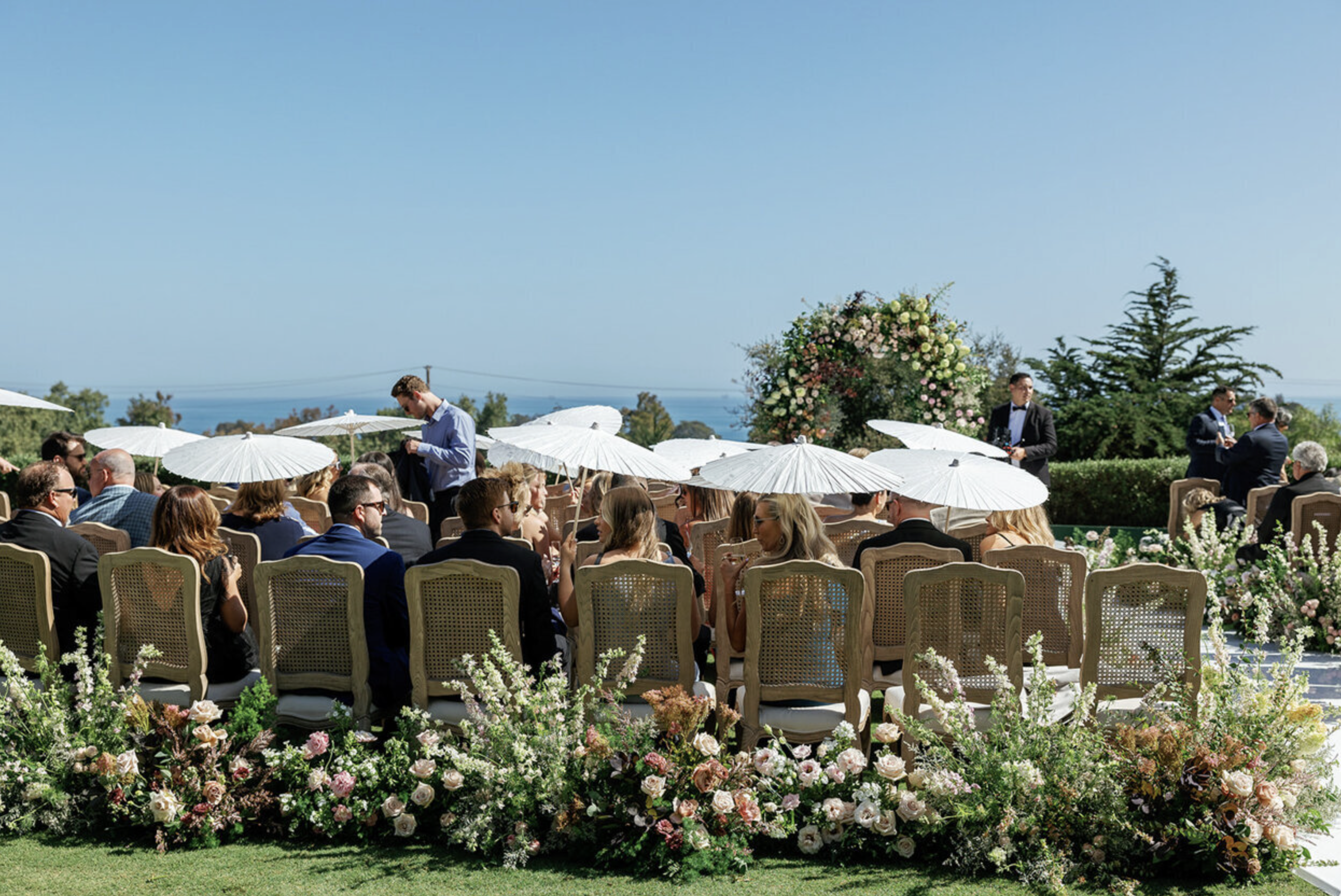 Wedding guests seated with white parasols at an outdoor ceremony overlooking the Pacific Ocean at a Southern California venue, with a garden floral arch and rattan chairs