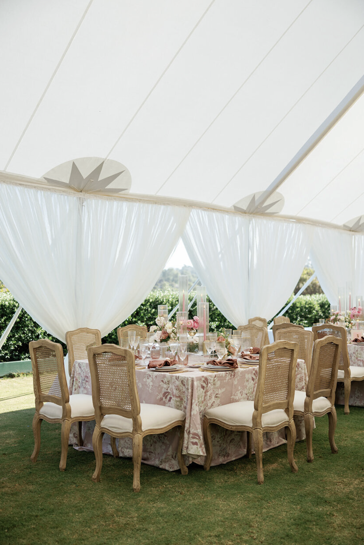 Tented outdoor wedding reception with round tables, floral pink linen tablecloths, rattan cane chairs, and pink taper candles, styled by a California wedding producer