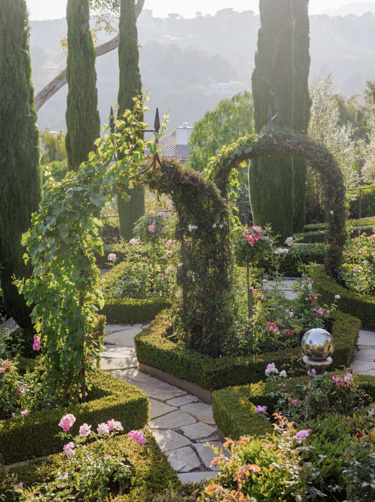 Rose garden with vine archway, manicured boxwood hedges, and cypress trees at Villa Sancti in Malibu, with hillside views in the background.