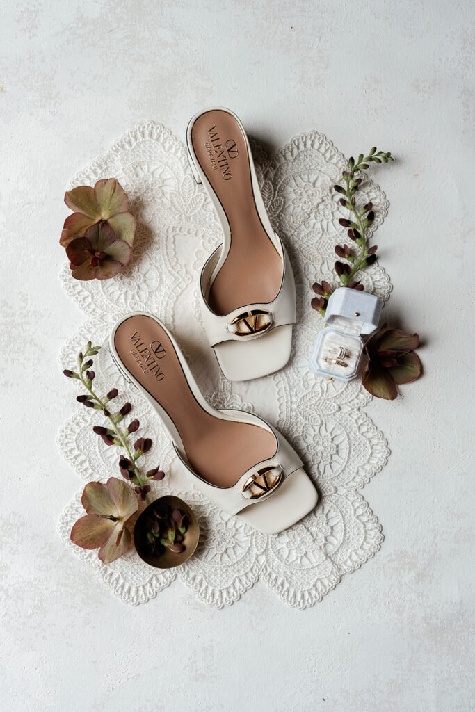Flatlay of ivory designer bridal heels and a wedding ring set in a pale blue ring box on lace, styled with dried hydrangea blooms, the kind of detail shot a planner styles before the day begins