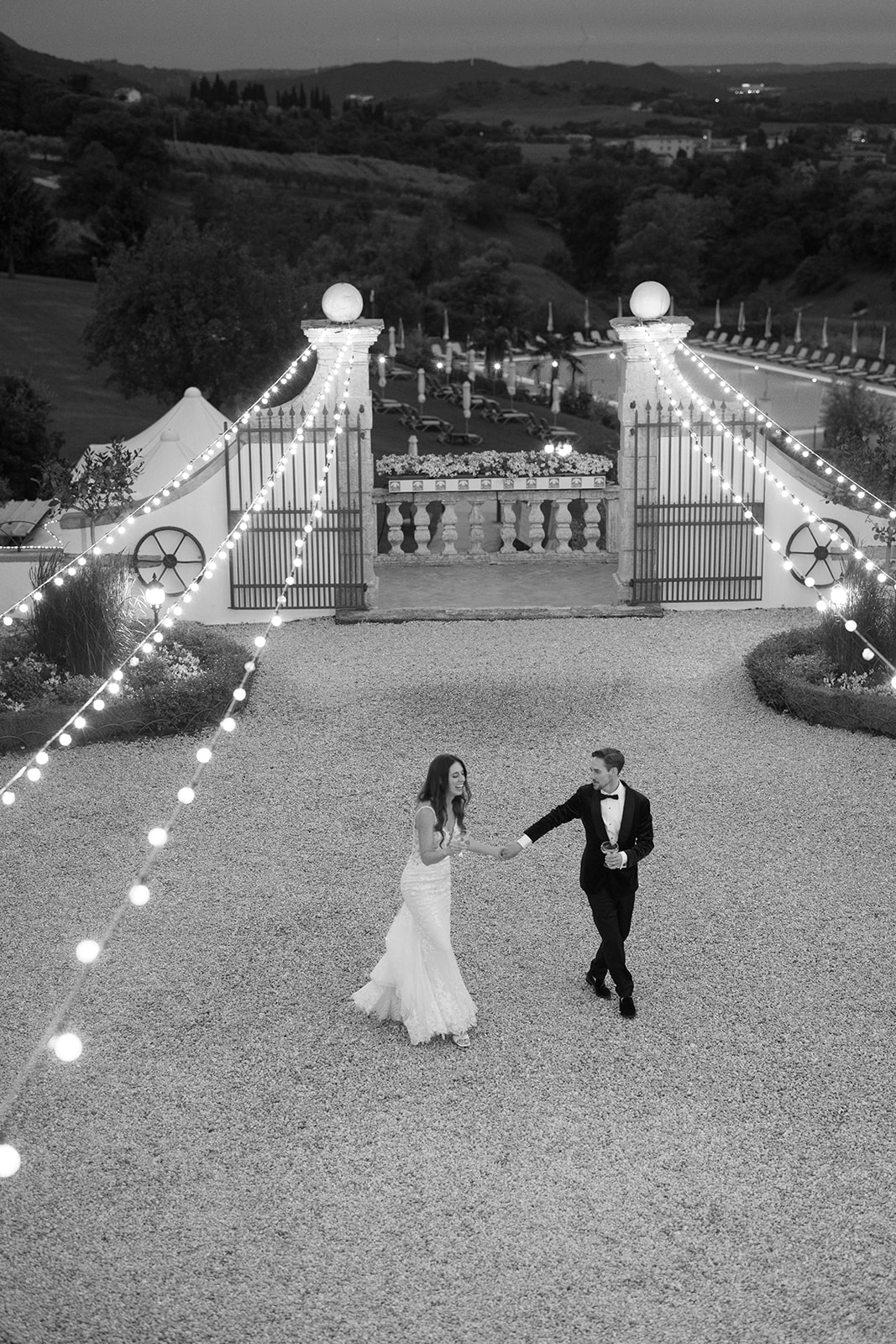 Black and white photo of a bride and groom holding hands in a gravel courtyard under string lights with rolling hills behind them at a destination wedding