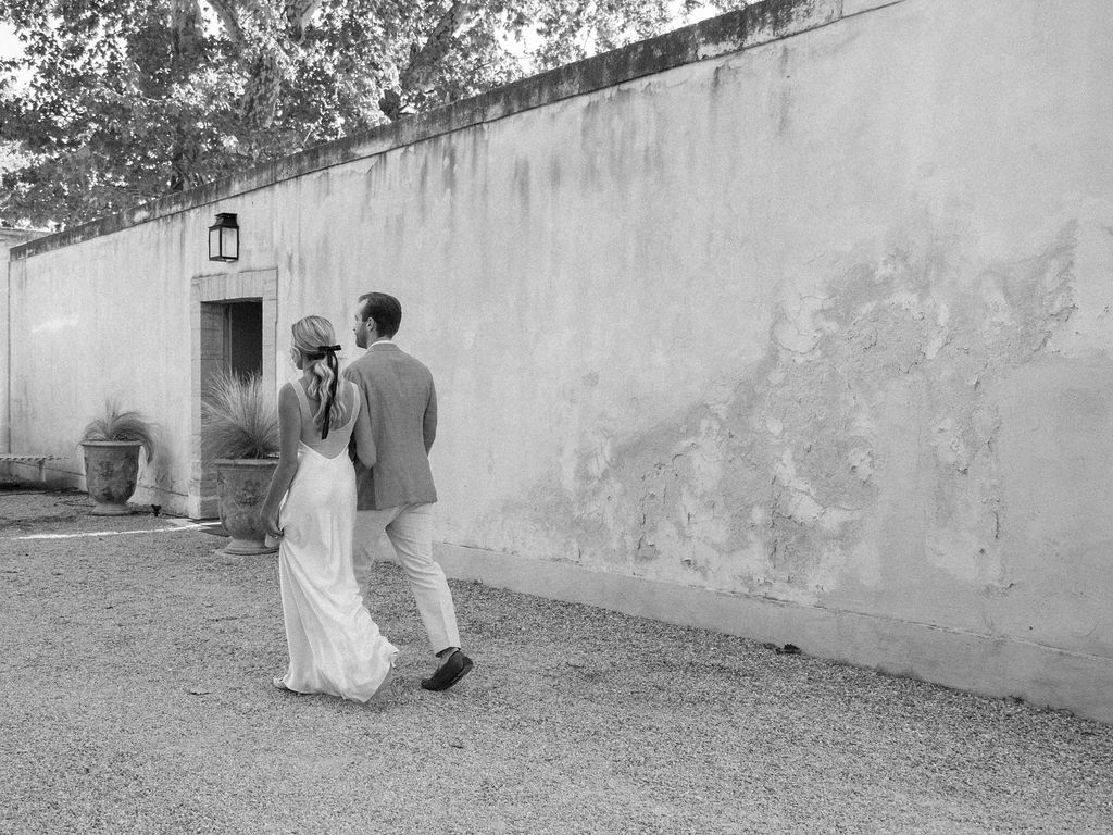 Black and white photo of a bride and groom walking arm in arm along a weathered stone wall at a European wedding venue